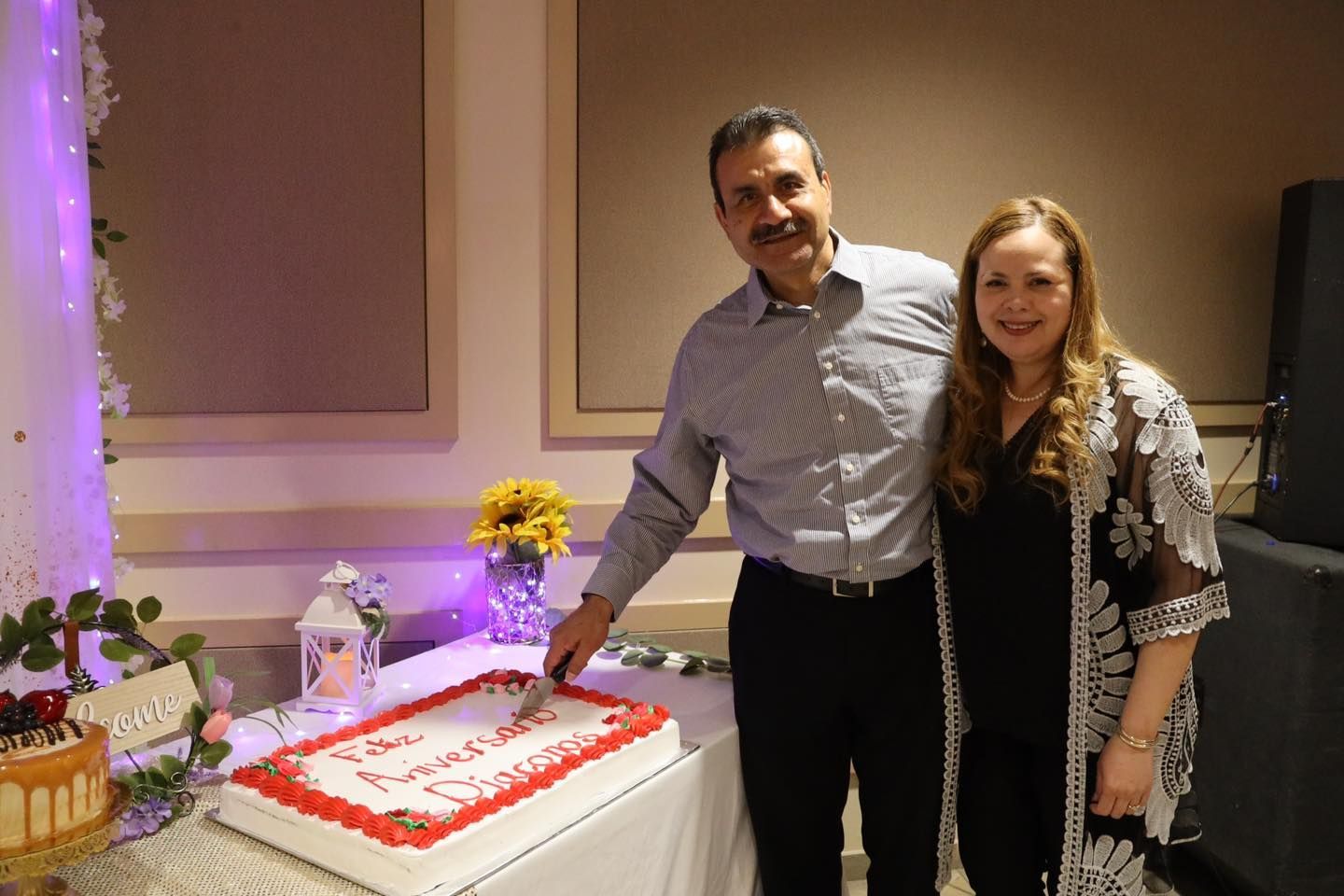 Two people standing beside a decorated cake at a celebration table indoors.