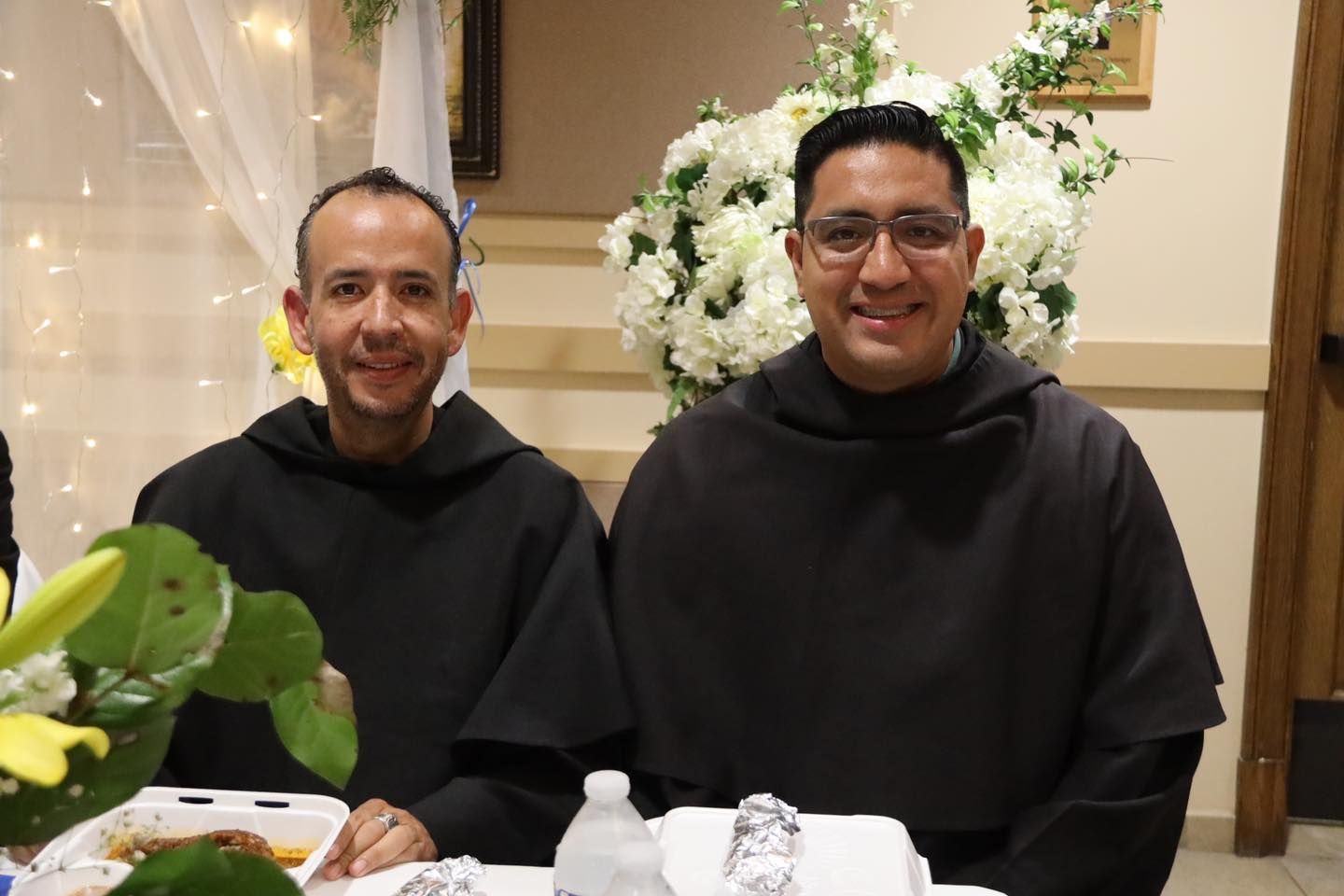Two smiling monks seated at a banquet table with white flowers and warm decorative lights behind them
