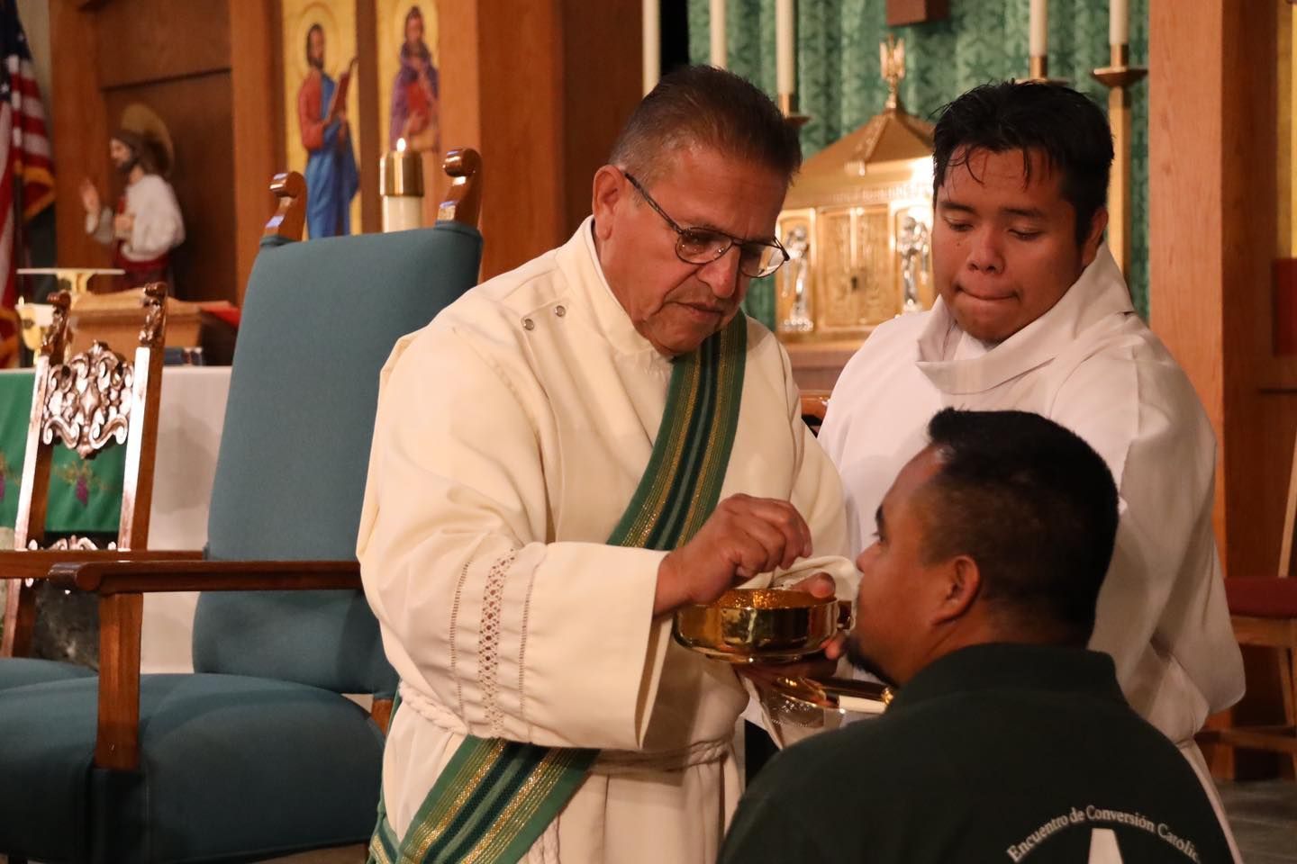 Priest giving communion to a seated person in a church, with an assistant standing beside him.