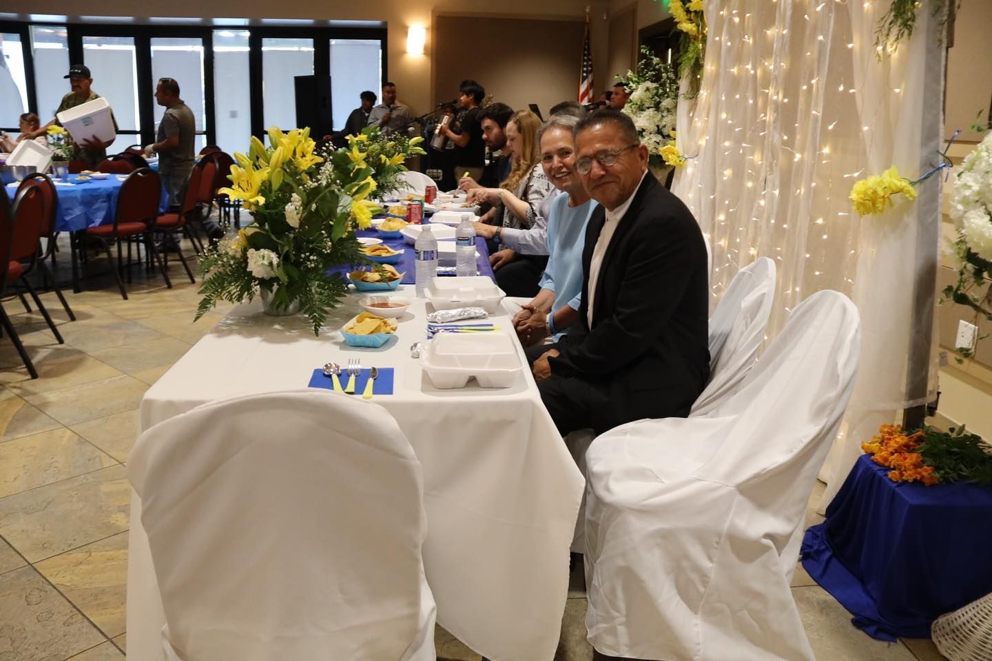 Formal banquet table with seated guests, floral centerpieces, and white drapery in a decorated hall