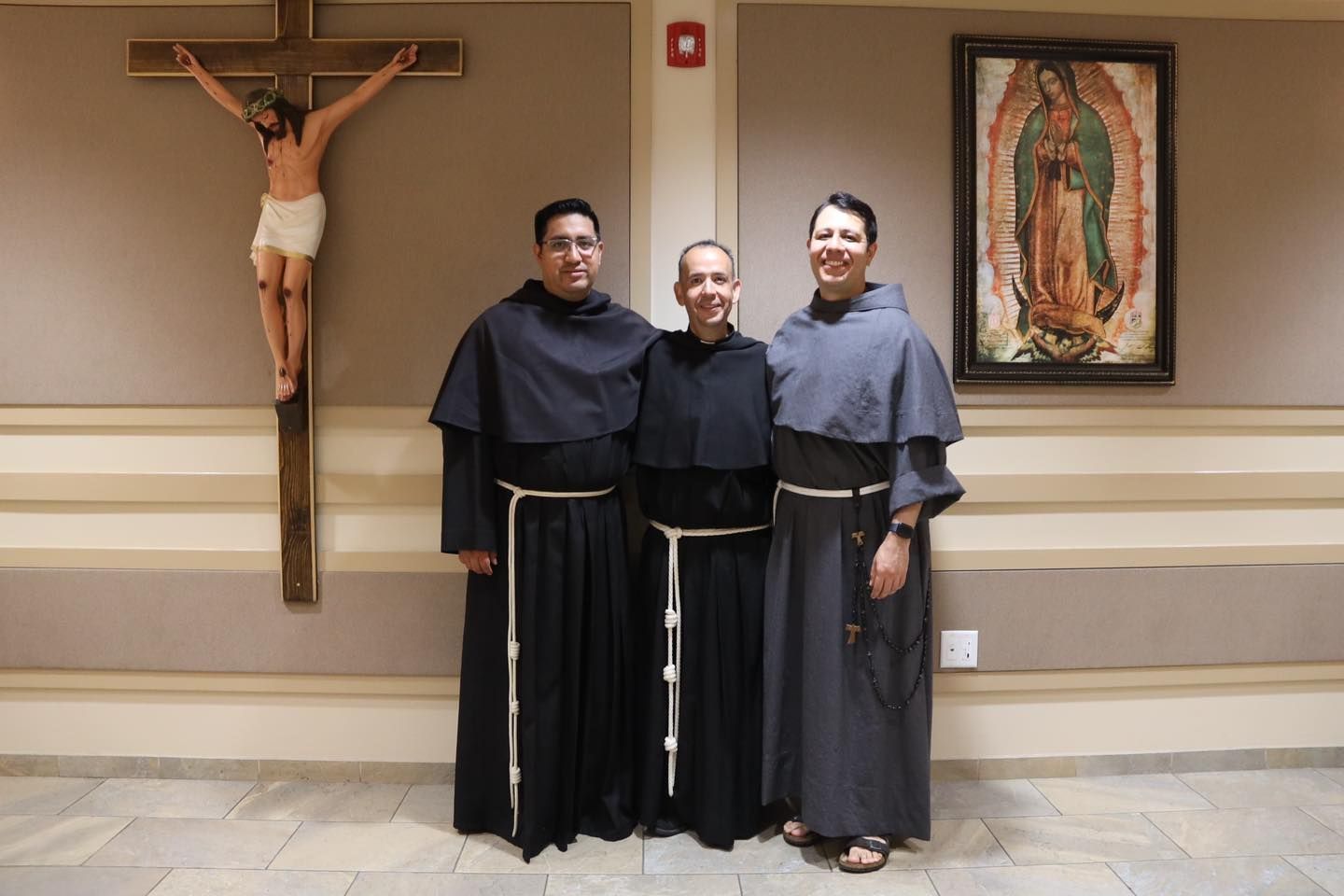 Three people in religious robes posing beside a crucifix and Our Lady of Guadalupe painting.