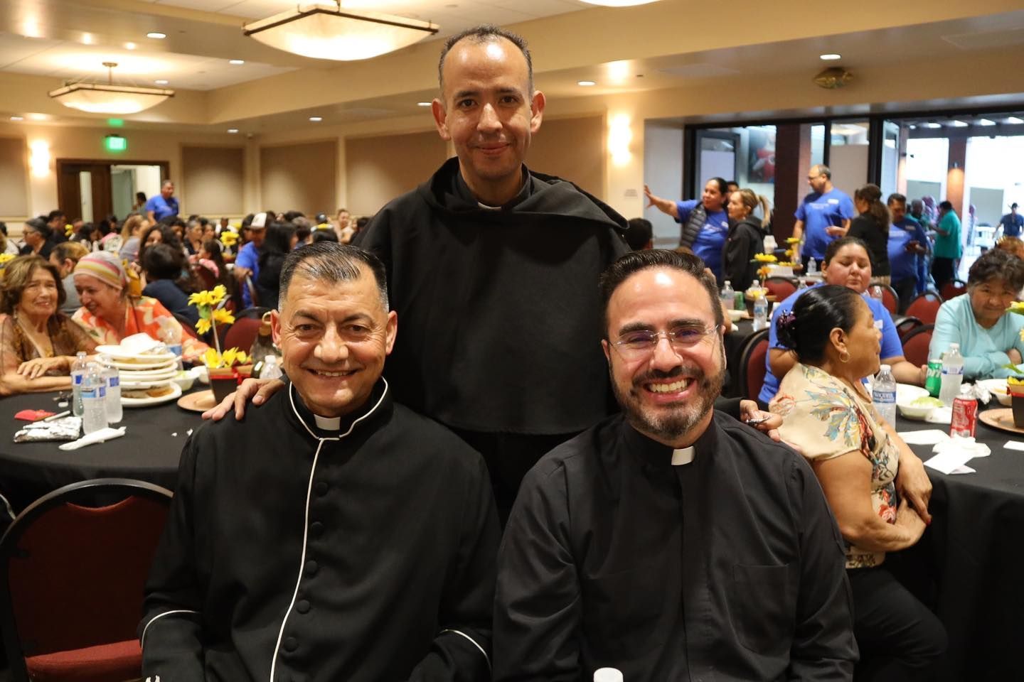 Three men in black clerical attire smiling at a banquet table in a crowded hall