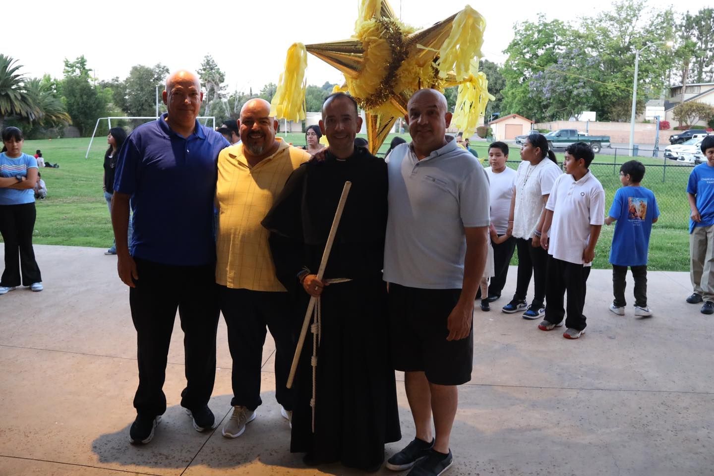Four adults pose with a large gold star balloon; children stand in line behind them outdoors.