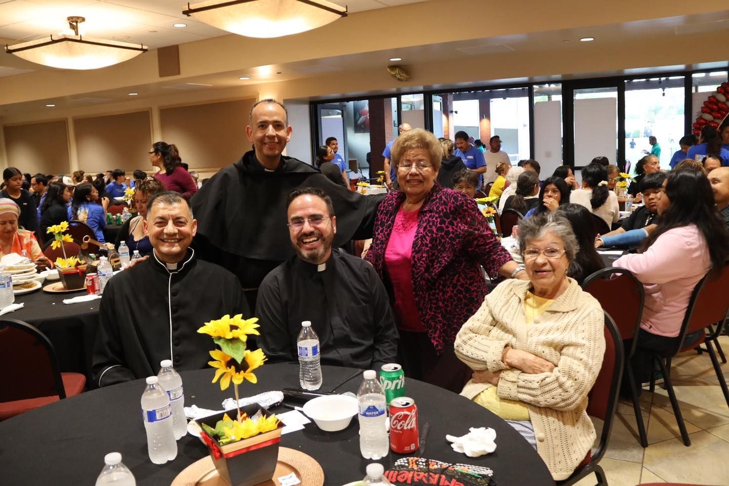 People seated at banquet tables in a community hall, with four smiling guests in the foreground.