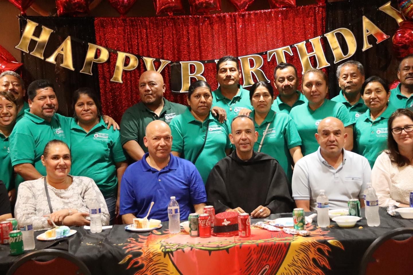 Group of people at a birthday celebration seated before a “Happy Birthday” banner and decorated table