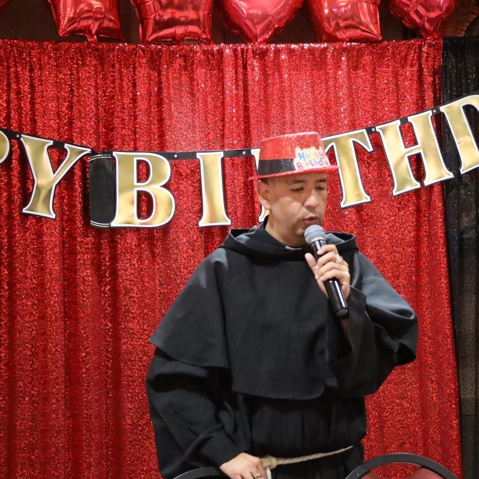 Person speaking into a microphone on a red-decorated stage with a “BIRTH…” banner behind them
