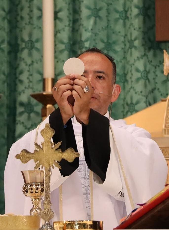 Priest holds up a communion wafer during Mass beside a gold cross and candle altar decor
