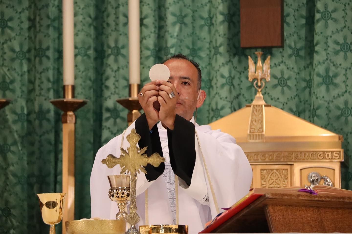 Priest elevates a communion wafer before a gold altar during Mass