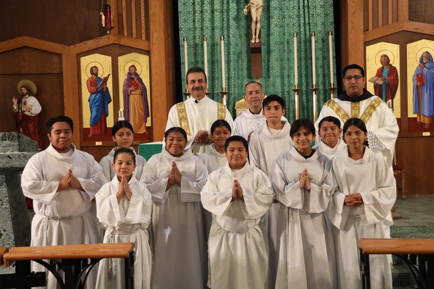 Group of altar servers in white robes standing in a church with hands folded in prayer.