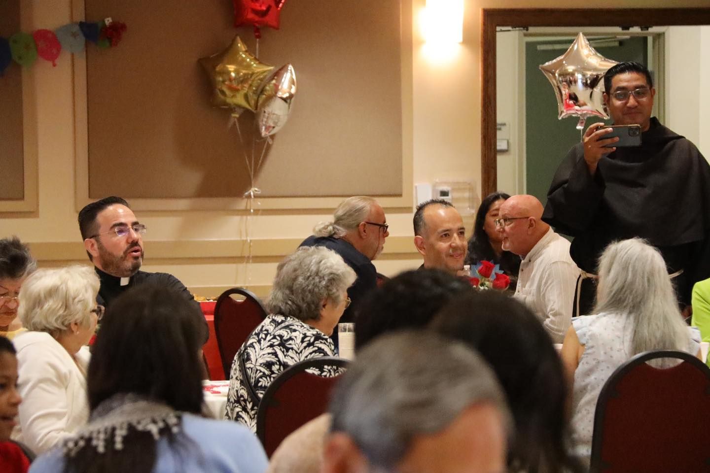 Man speaking into a microphone at a crowded indoor gathering with seated guests and decorations