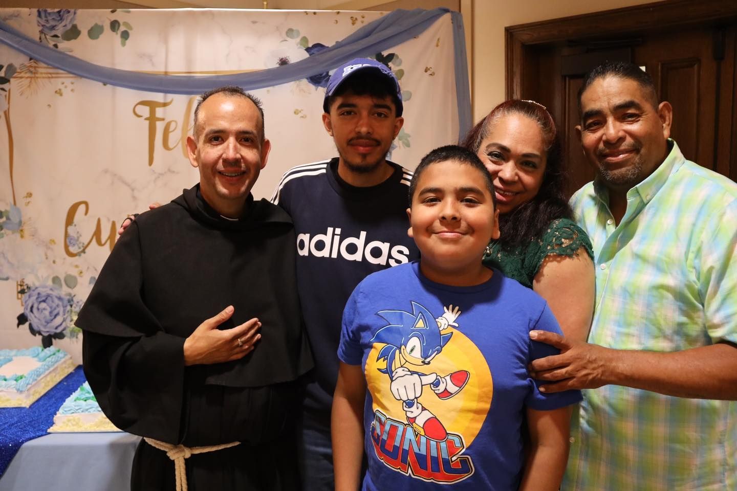 Five people smiling indoors at a celebration, with a decorative banner in the background.