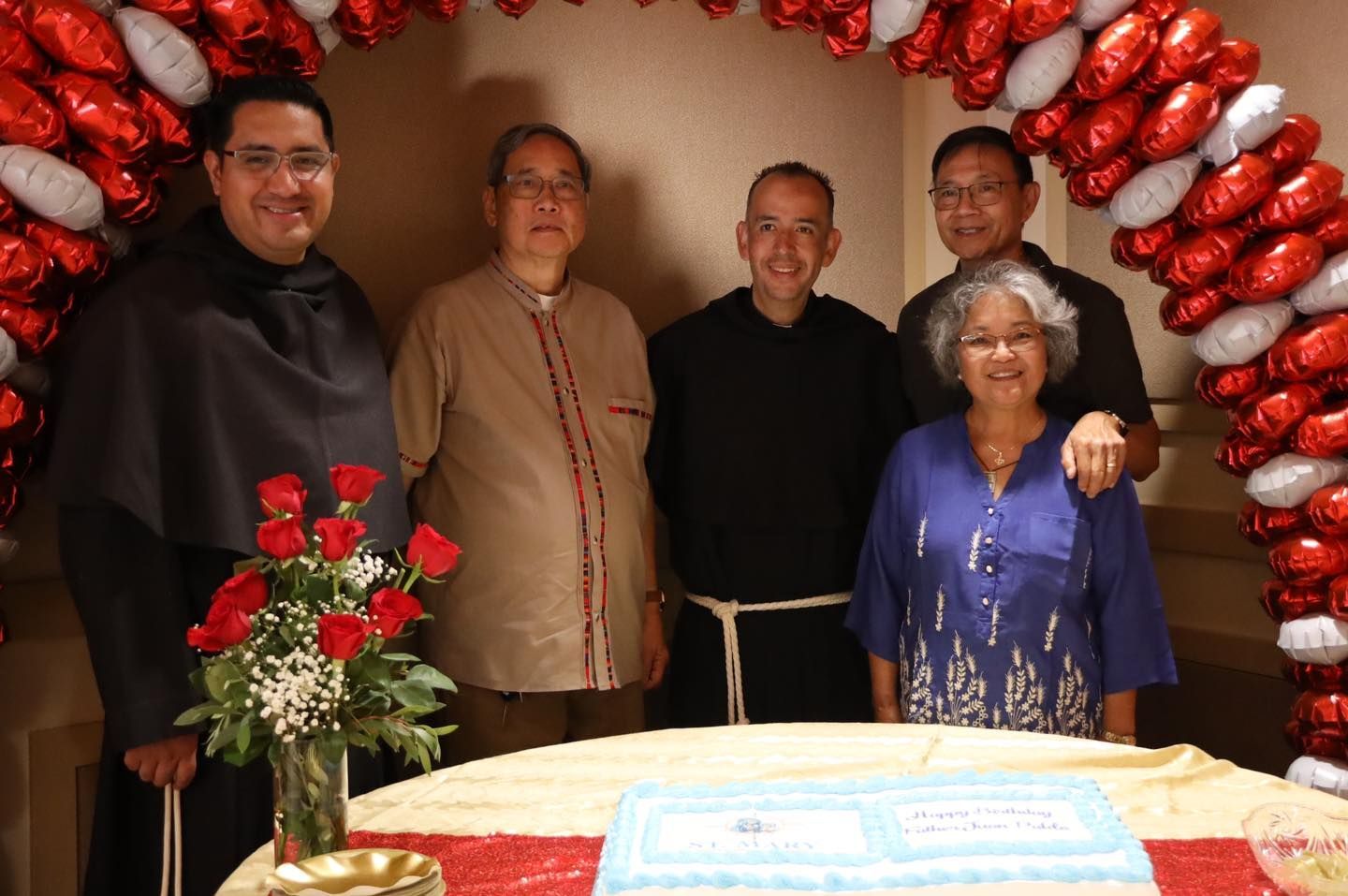 Five people standing behind a decorated cake and red rose bouquet in a festive indoor setting