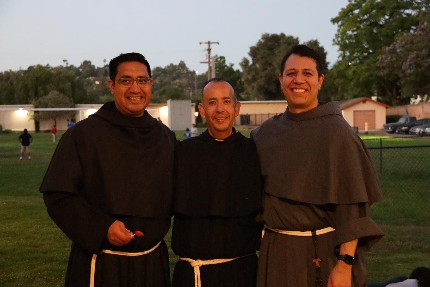 Three smiling people in dark robes standing outdoors at dusk on a grassy field.