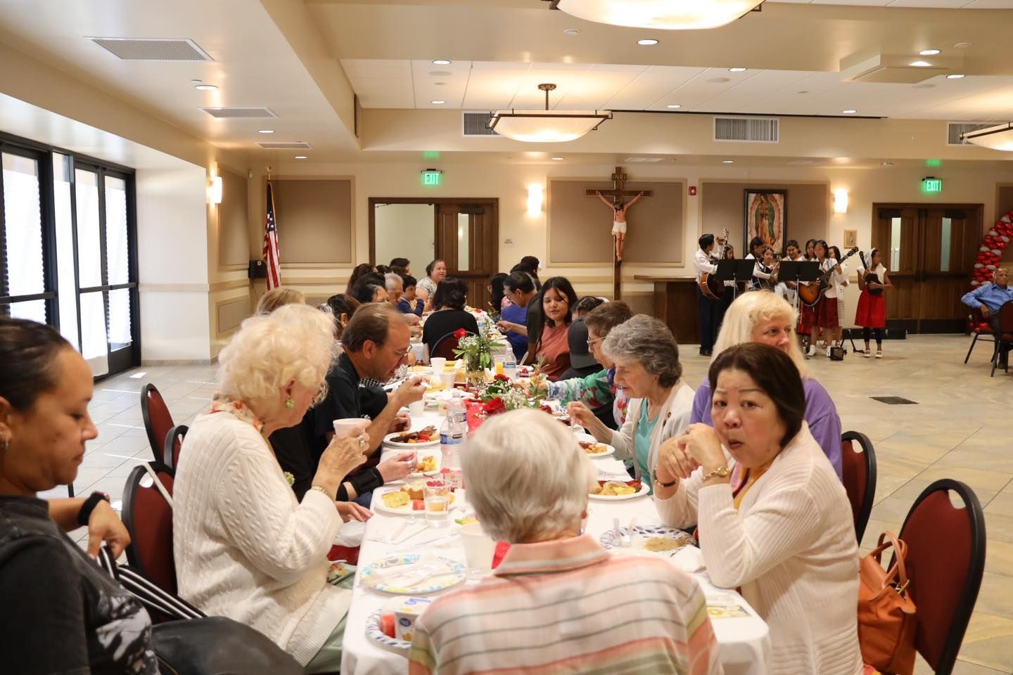 People seated at long tables in a community hall, chatting during a shared meal event.