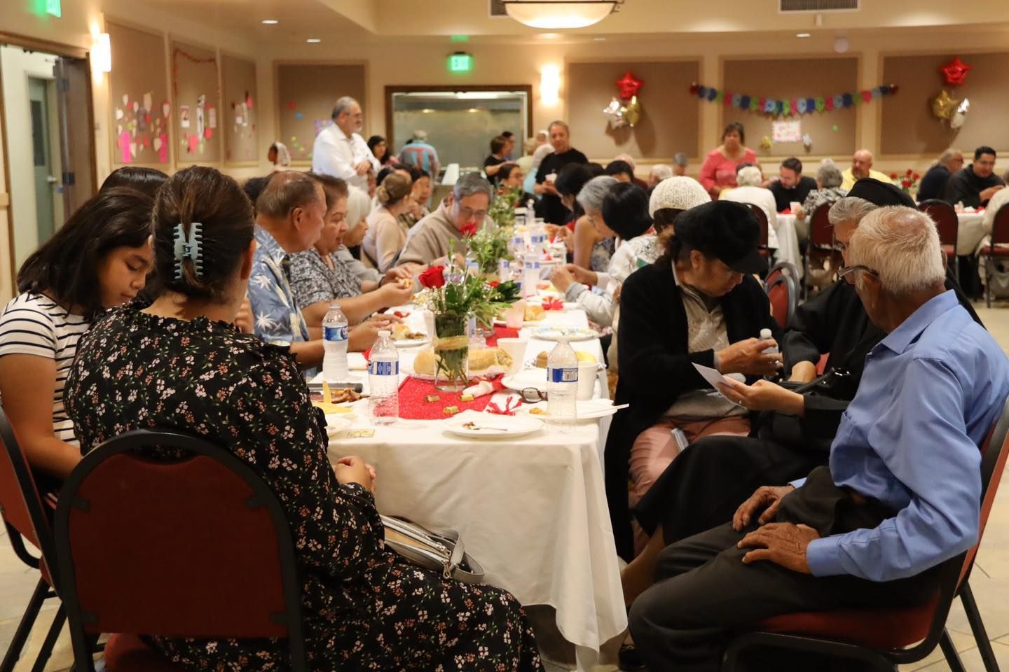 Banquet guests seated at decorated tables in a warmly lit community hall, eating and chatting