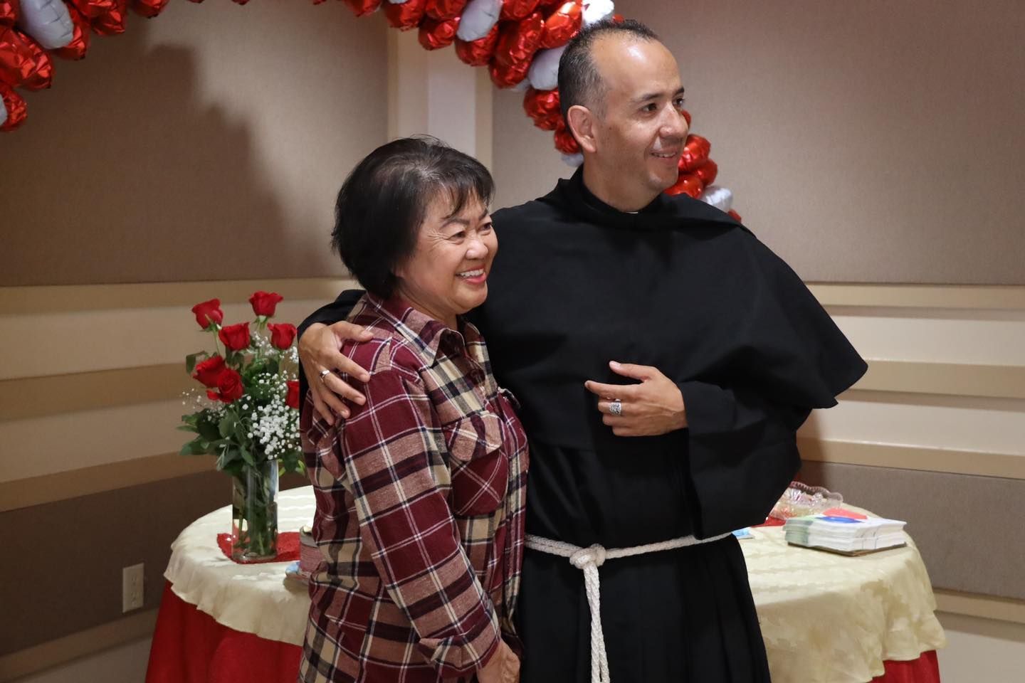 Two people smiling and posing indoors beside a decorated table with red roses and garland