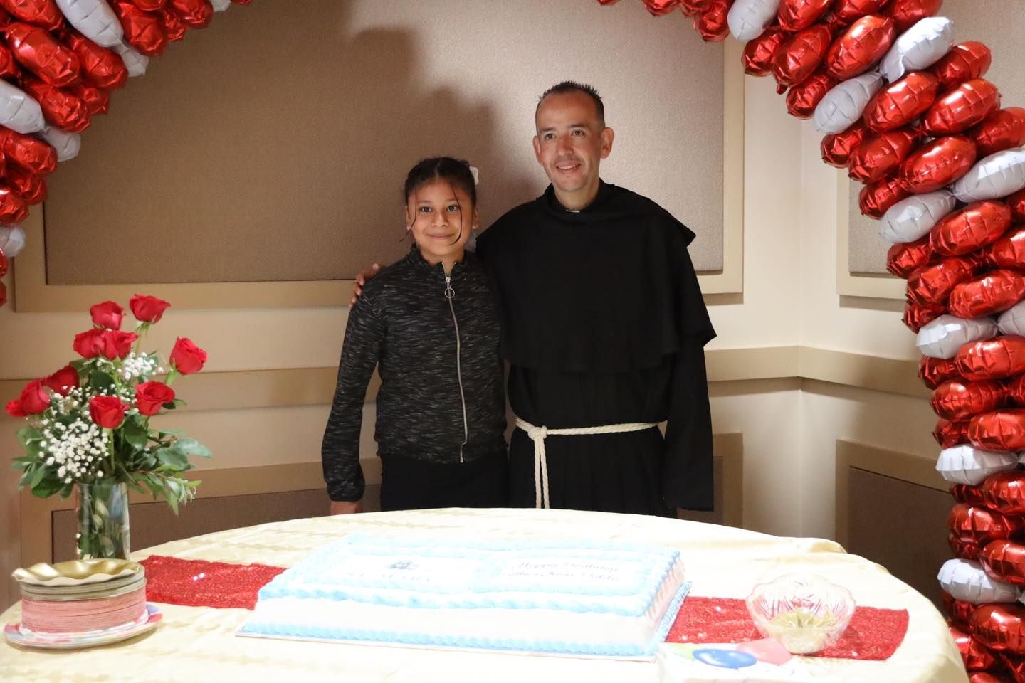 Two people standing behind a decorated table under a red-and-white balloon arch