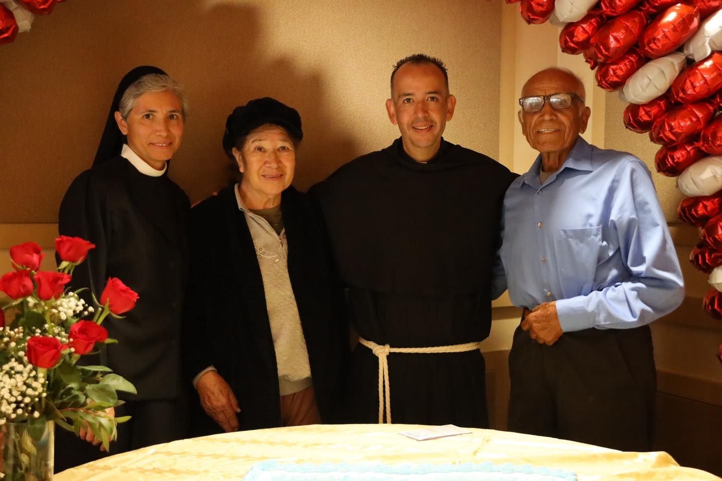 Four people standing together indoors, smiling beside a table with flowers on the left.