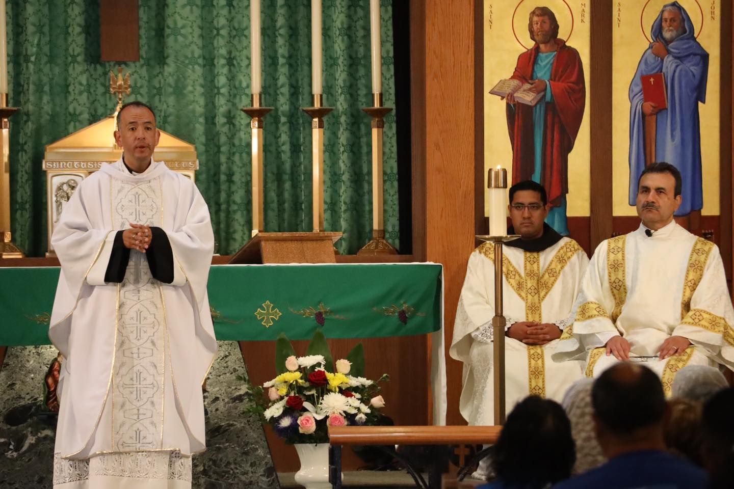 Priest standing at church altar with two seated clergy behind him and religious icons in the background