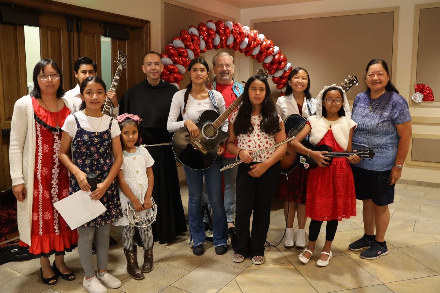 Group posing indoors with red-and-white balloon arch, holding bouquets and a saxophone