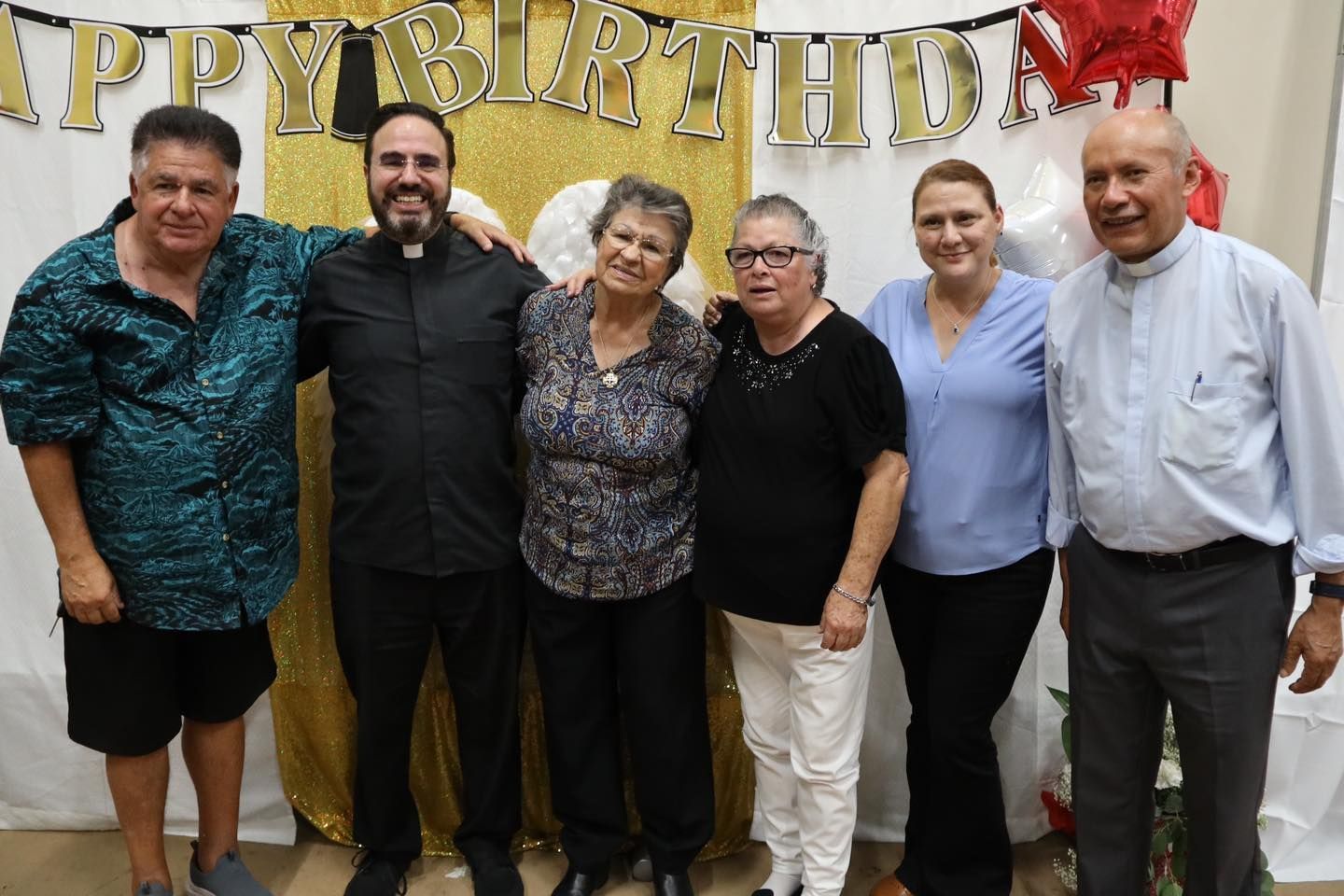 Six people posing at a birthday party in front of a gold “Happy Birthday” banner.