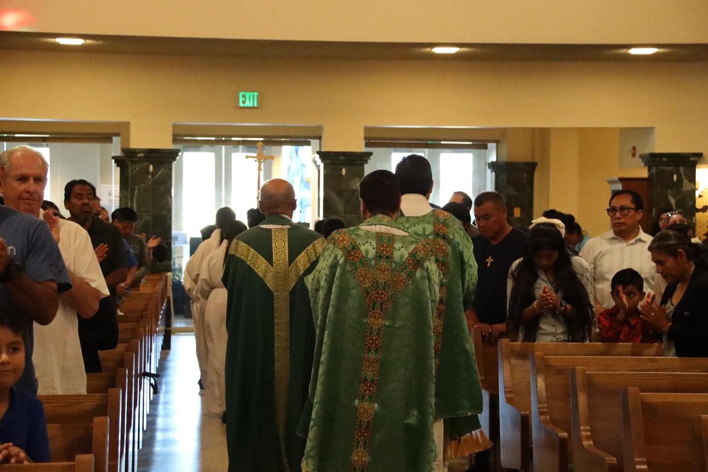 Priests in green vestments process down a church aisle during a service, with seated congregants on both sides.