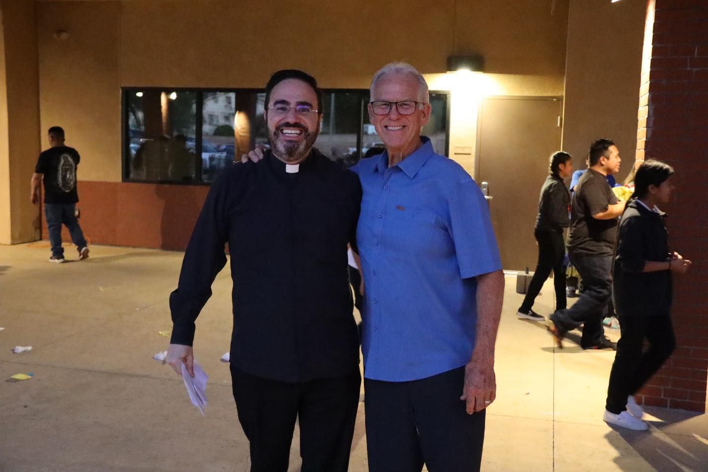 Two smiling men stand together outside a building at night, with people walking in the background.