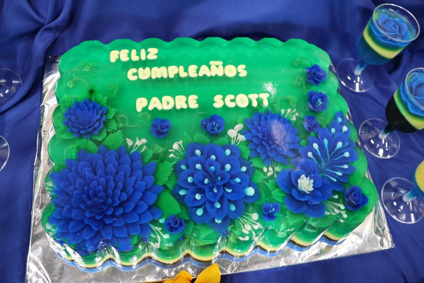 Birthday cake with blue flowers and “Feliz cumpleaños Padre Scott” written in white icing.