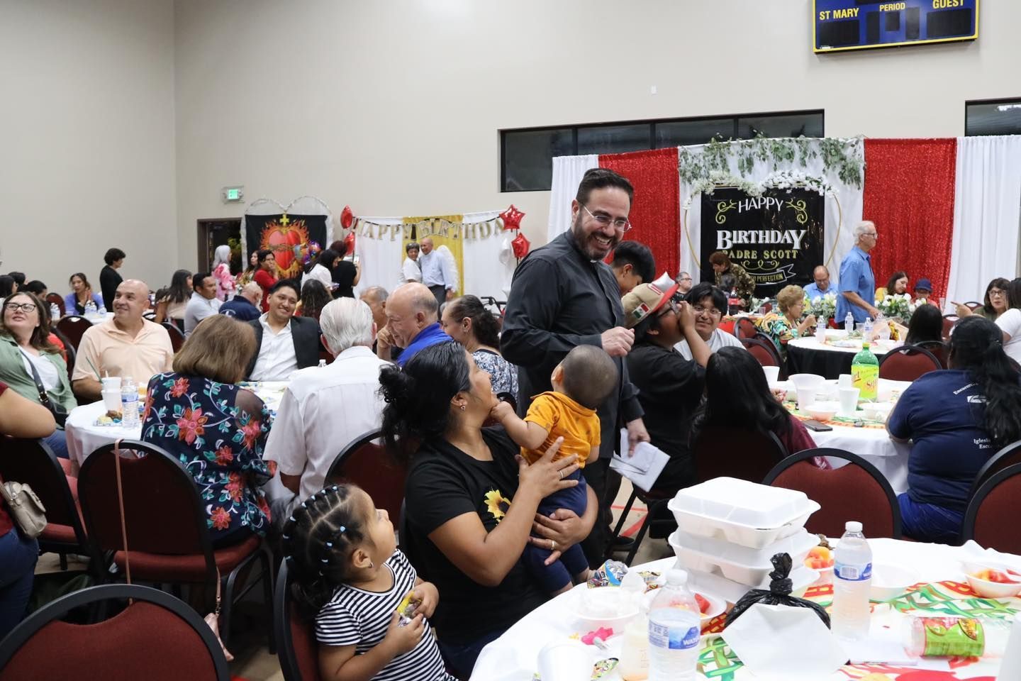 People seated at a banquet hall event, with a man standing and speaking near decorated tables.