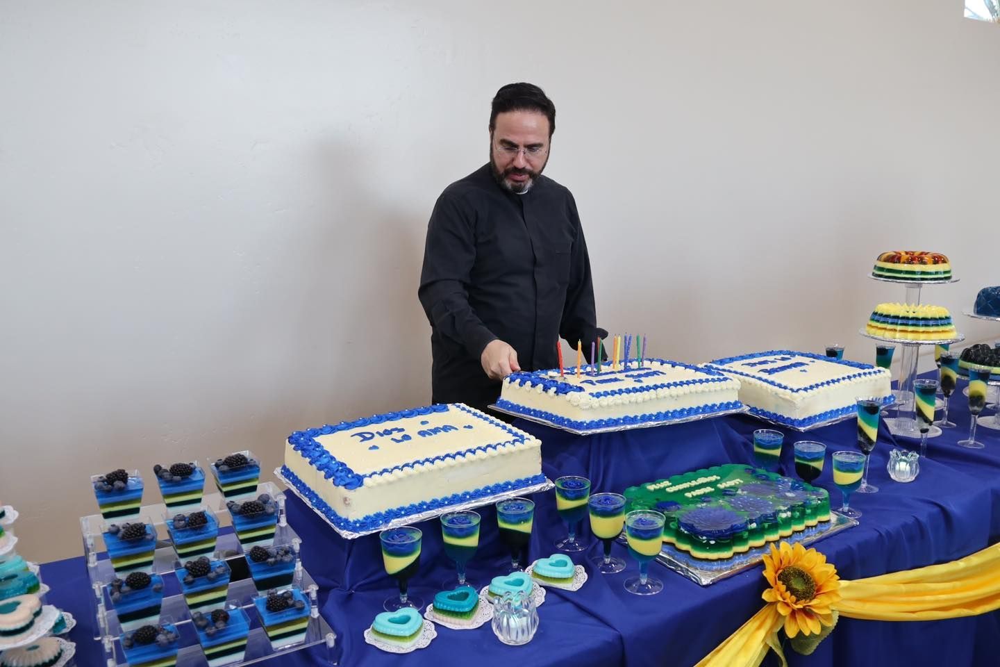 Person standing behind a blue-and-white decorated cake display on a table with cupcakes and a sunflower centerpiece