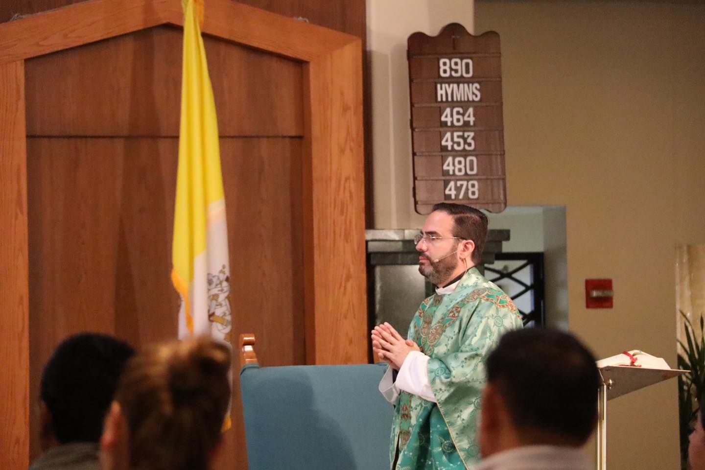 Priest in green vestments speaking at a church lectern with congregation seated in foreground