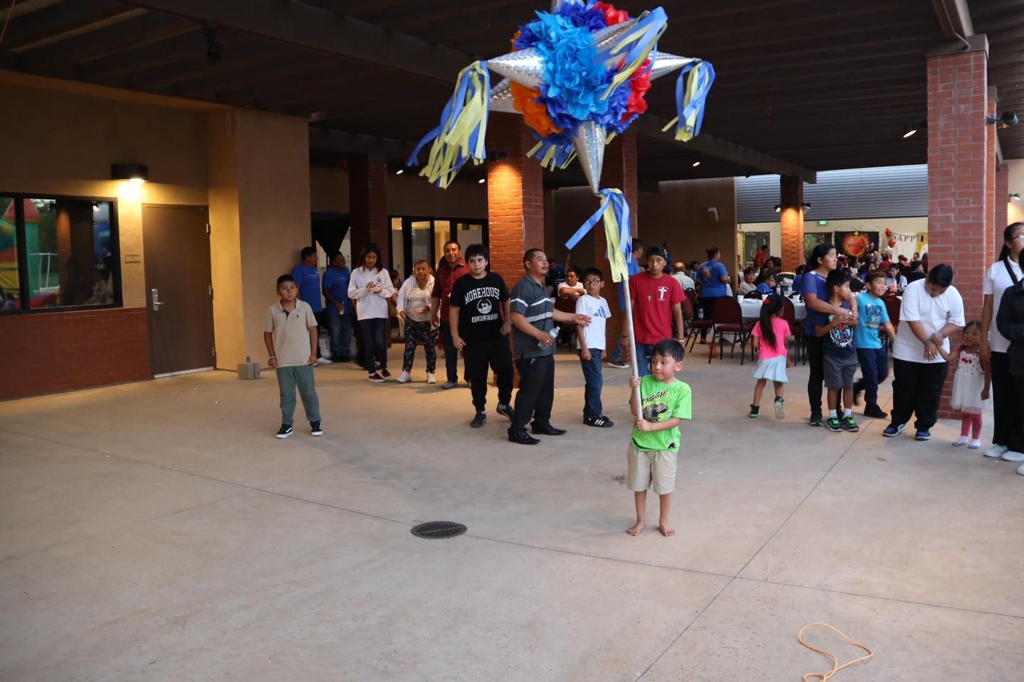 People gathered under a building overhang beside a colorful hanging decoration, with a child in green in front.