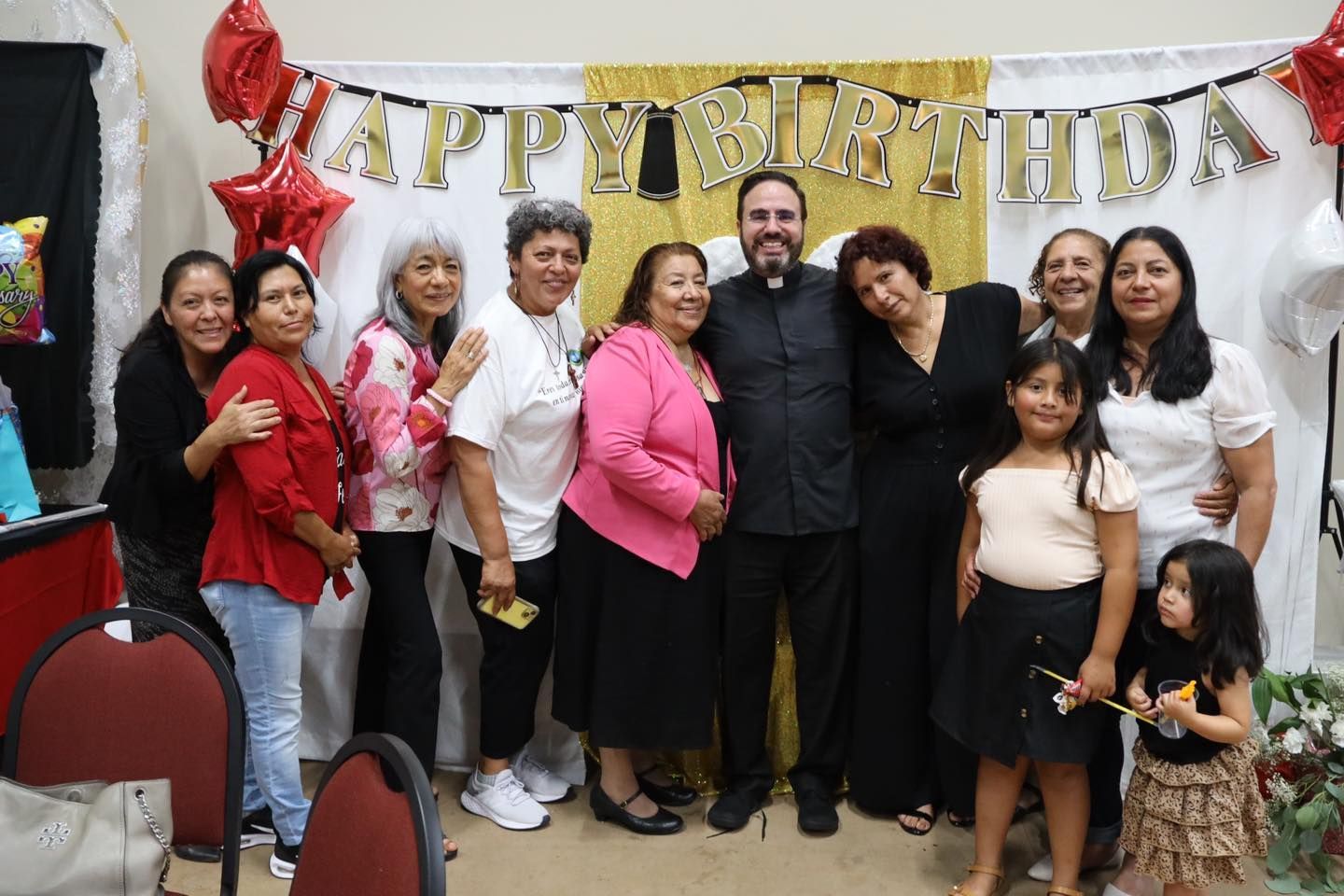 Group posing at a birthday party with gold “Happy Birthday” banner and red decorations
