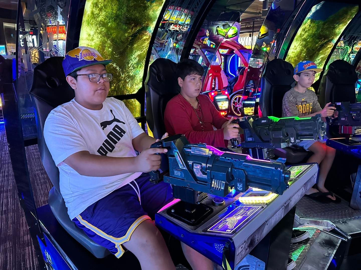 People seated at arcade racing games, focused on steering neon-lit consoles in a dark arcade.