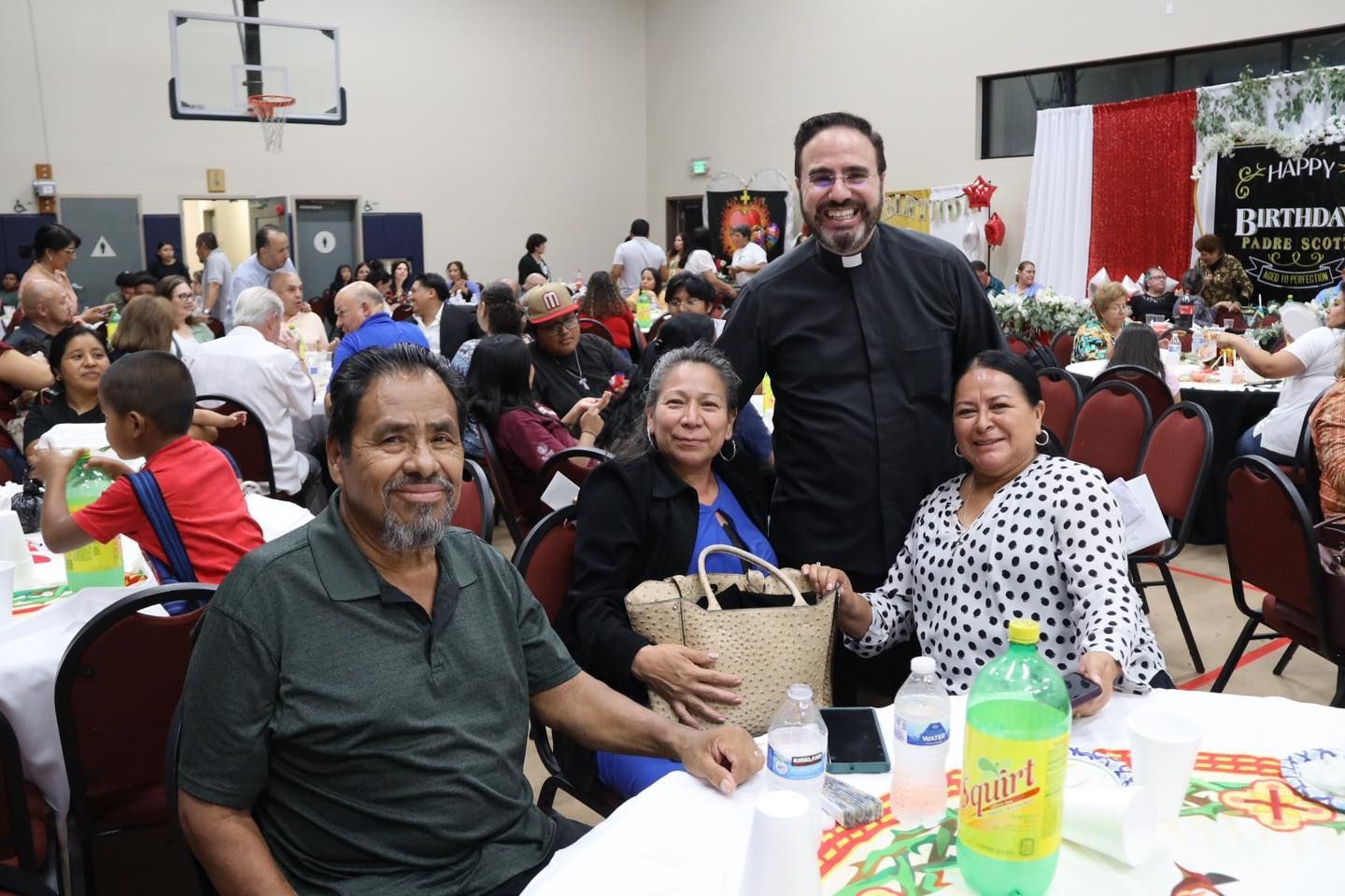 Group seated at a banquet table in a busy hall, with a man standing behind them smiling.