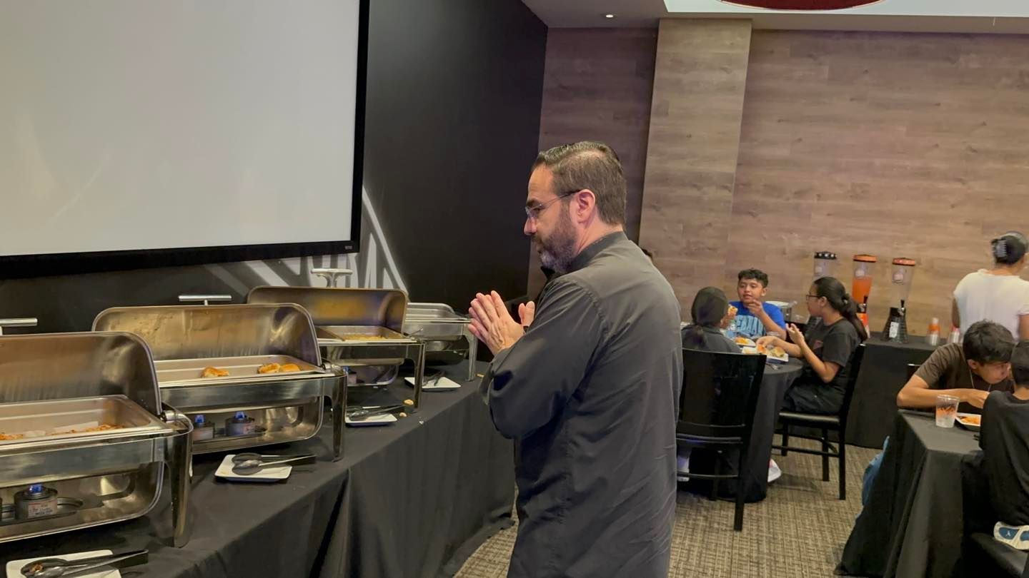 Man clapping beside a buffet table in a banquet hall while attendees sit at tables in the background