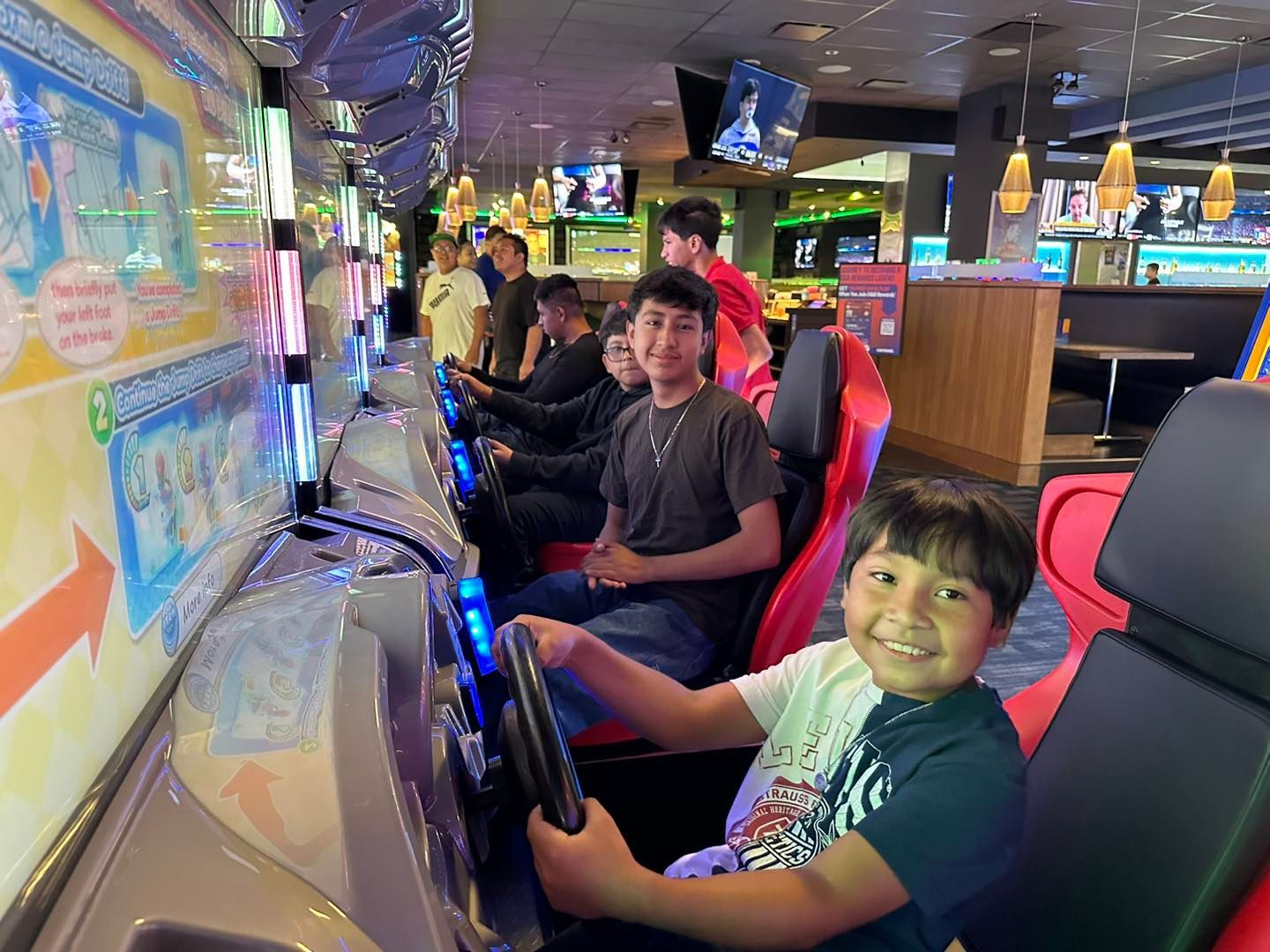 People playing arcade racing games in a colorful game room, with one smiling at the camera.