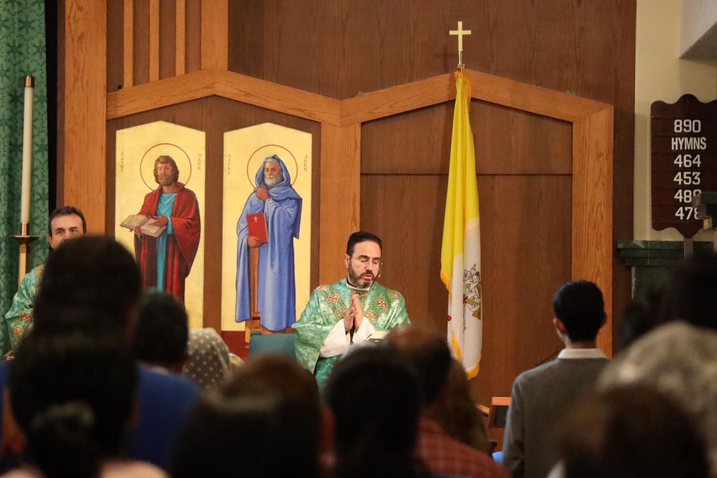 Priest in green vestments speaking at church altar before icons, with congregation standing in foreground