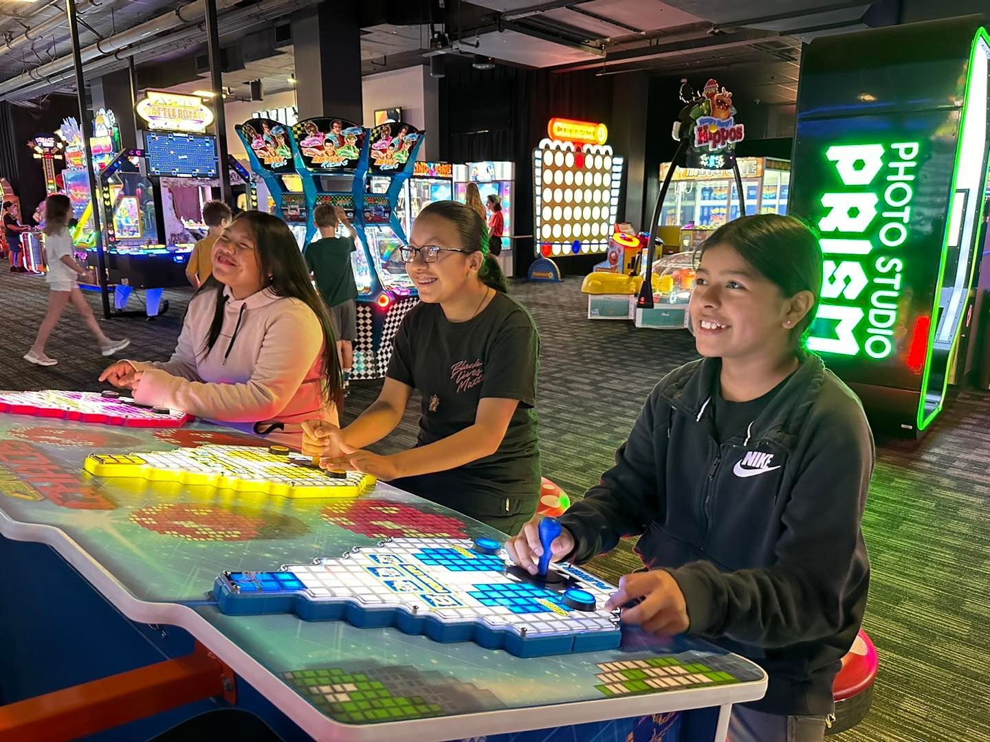 Three people play a glowing carnival arcade game under neon lights, with a bright “Prism” sign nearby.