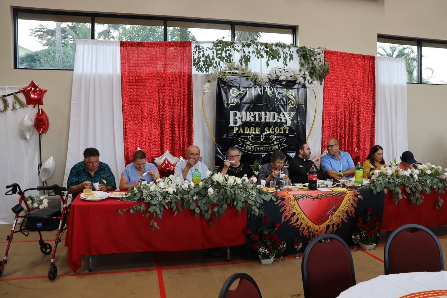 Formal panel seated behind red-draped table at decorated indoor event, with floral arrangement and banner backdrop