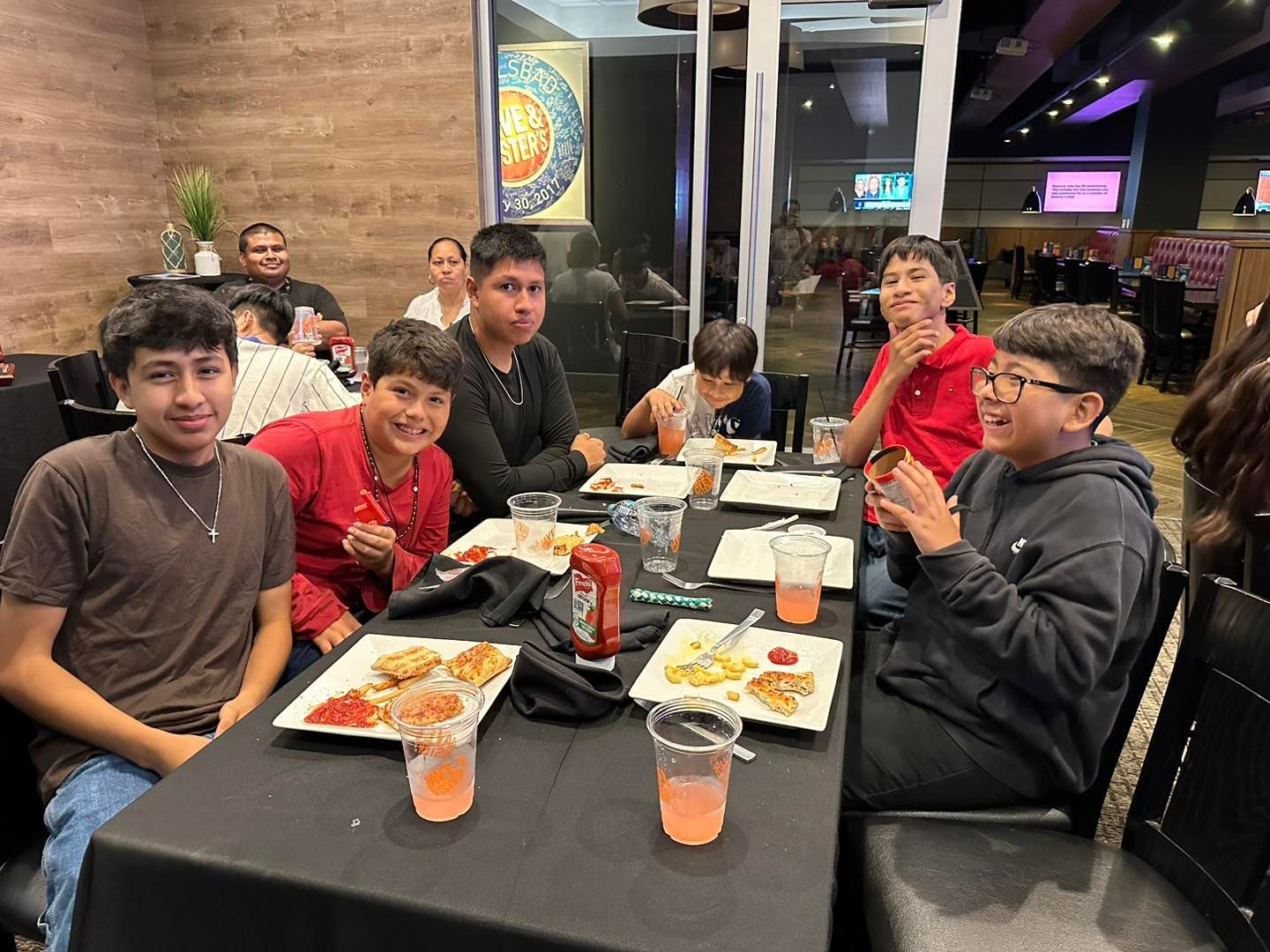 Group of six seated at a restaurant table, smiling and eating with drinks and plates in front of them.