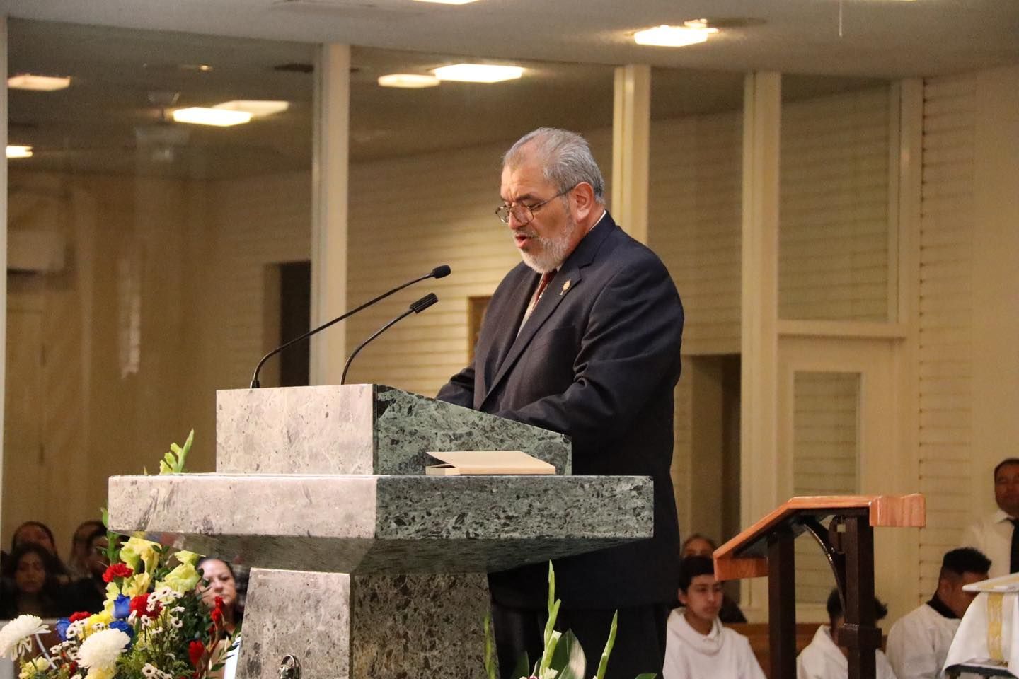 Man speaking at a marble podium in a church, with microphones and flowers in the foreground.