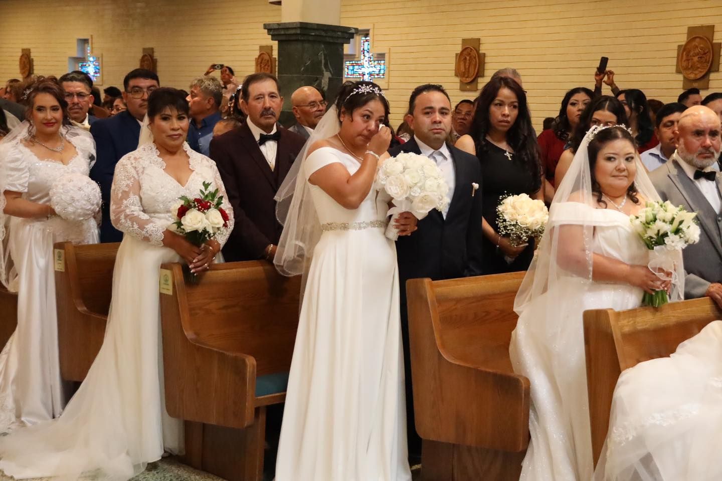 Brides and grooms standing in church pews during a wedding ceremony, holding bouquets and facing forward