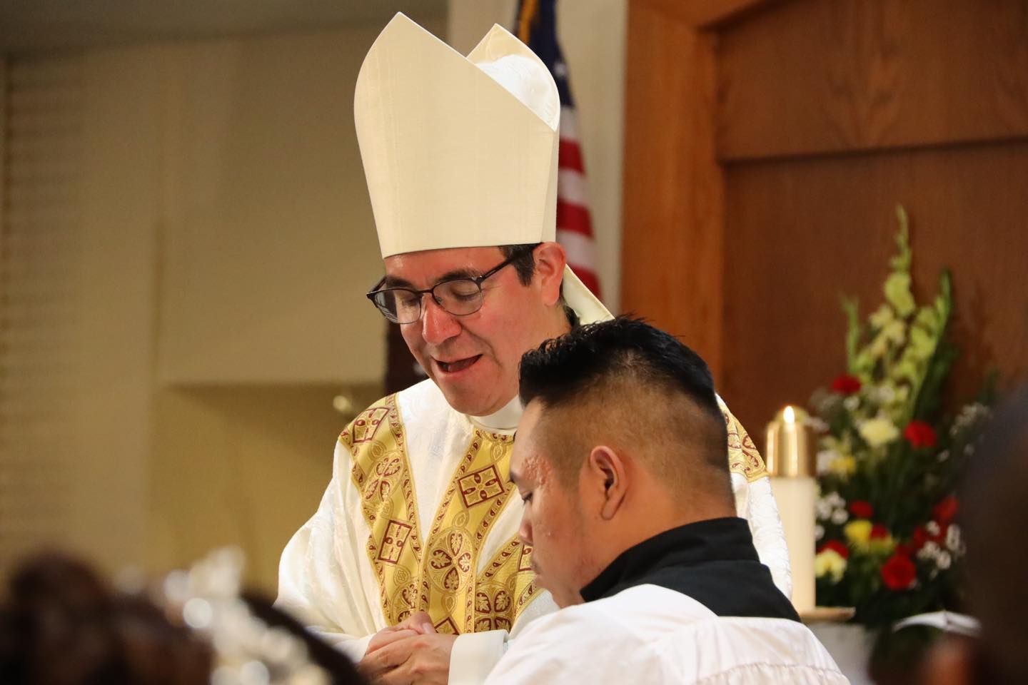 Priest in white and gold vestments speaking during a church ceremony, with an American flag behind him.