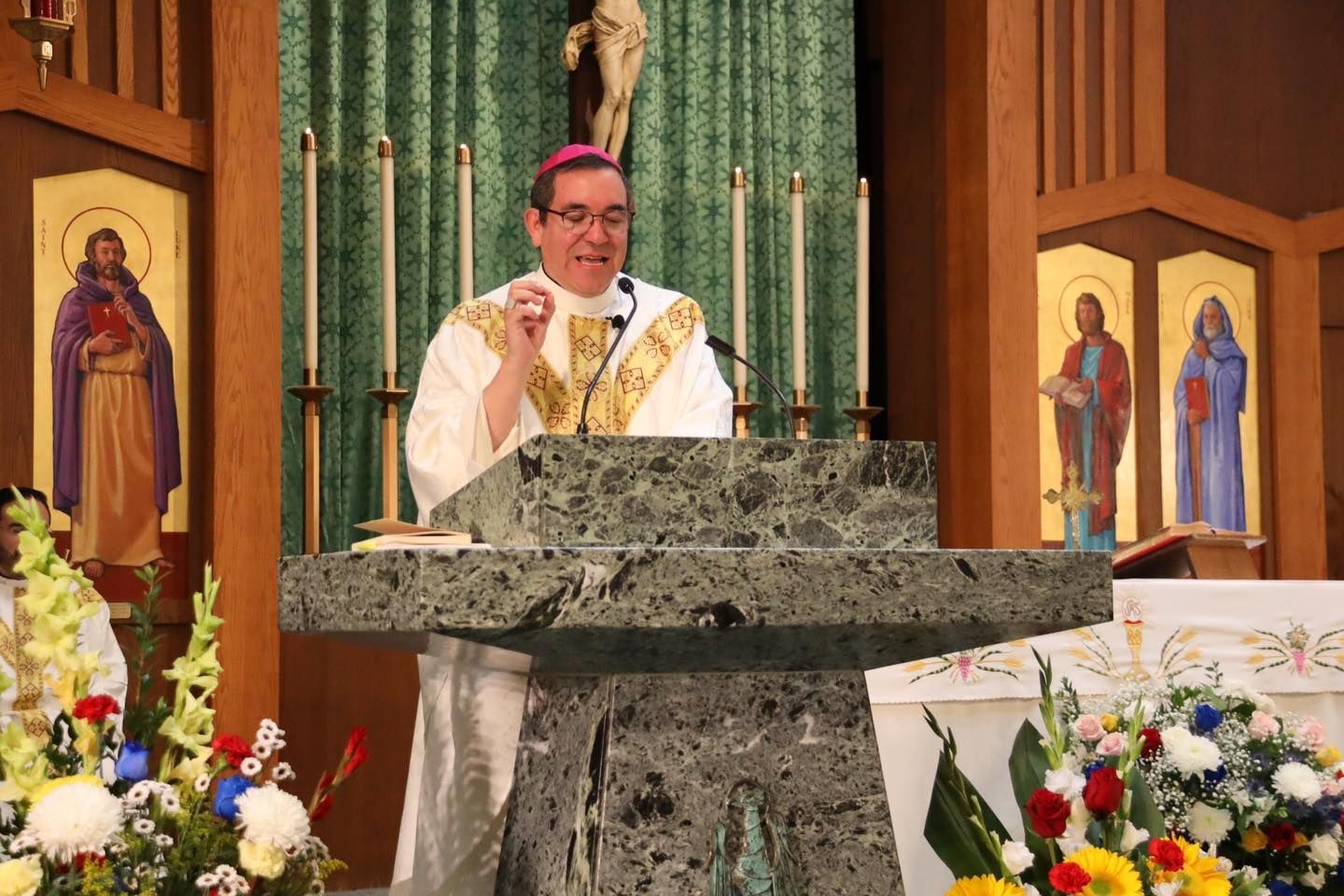 Priest speaking at an altar in a church, with floral arrangements and religious statues behind him