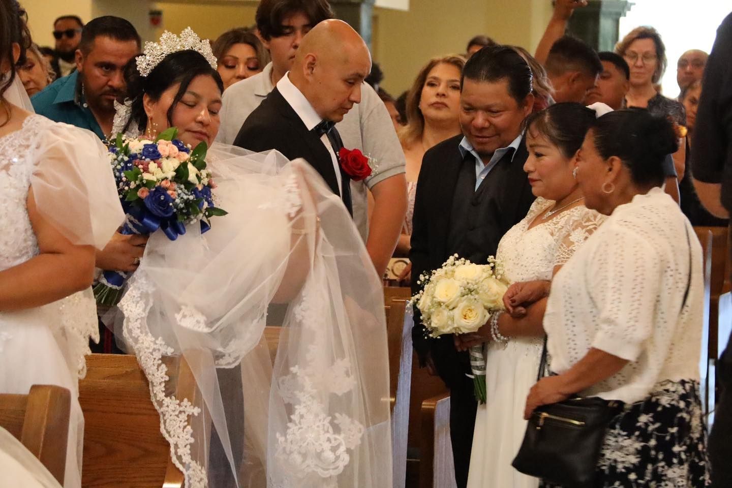 Wedding guests gathered indoors, with the bride holding a bouquet and a man exchanging flowers with another guest.