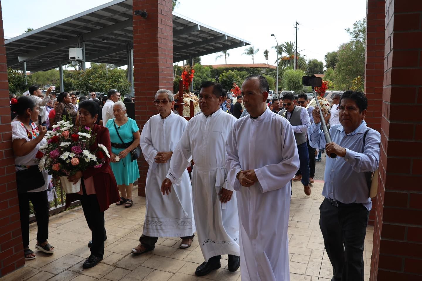 People in white robes enter a covered outdoor event, with flowers and onlookers nearby.