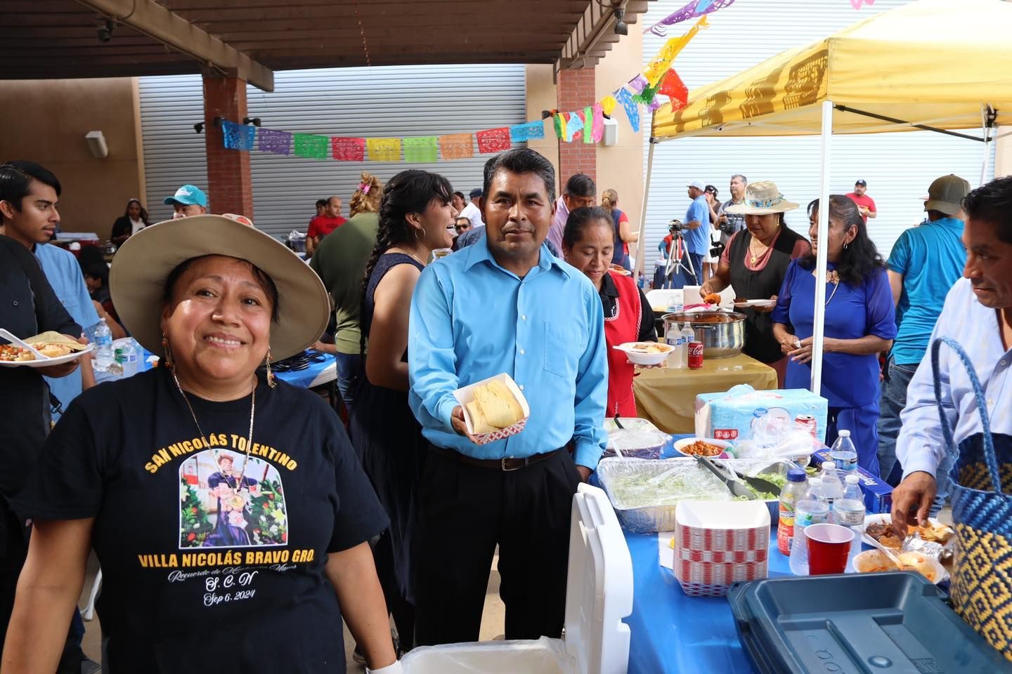 People at an outdoor market stall with colorful decorations and a man holding a coconut cake
