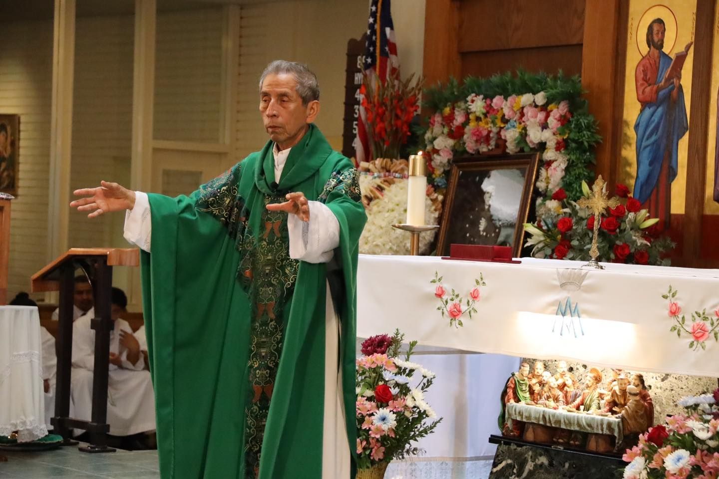 Priest in green vestments speaking before an altar decorated with flowers and religious icons