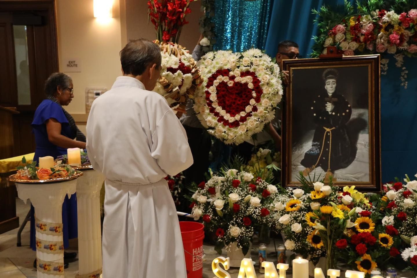Memorial altar with flowers, candles, and portrait; a person in white stands in quiet tribute.